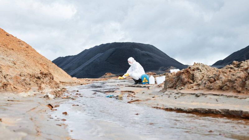 Taking sample of dirty water in toxic area Ecologist in white hazmat suit sitting at river and using pipette while taking sample of dirty water in toxic area